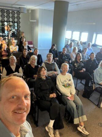 En man tar en selfie framför en grupp som sitter i ett rum