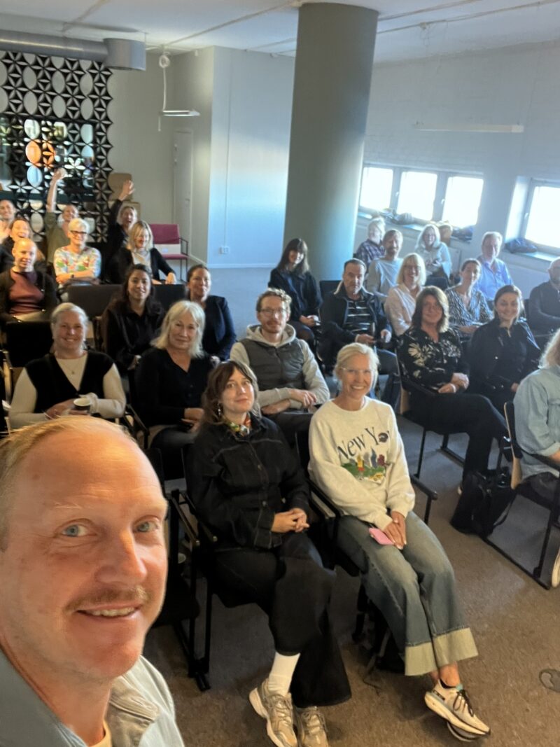 En man tar en selfie framför en grupp som sitter i ett rum